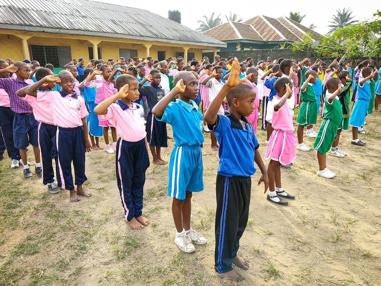 Group of schoolchildren in colorful uniforms saluting outdoors in a school yard.